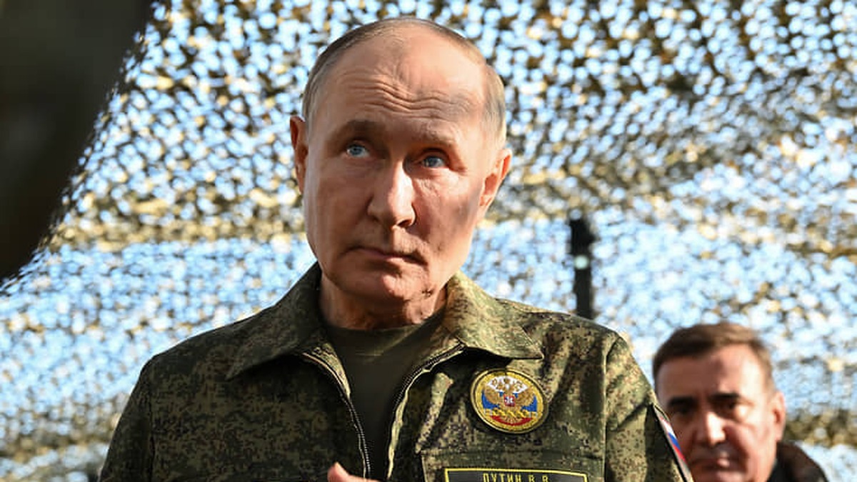 Vladimir Putin de uniforme militar camuflado sob uma rede de camuflagem, olhando para a frente